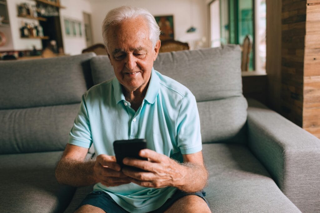 Mann ab 65 sitzt entspannt zuhause und lächelt auf sein Smartphone – erste Schritte ins Online-Dating nach dem Verlust des Partners