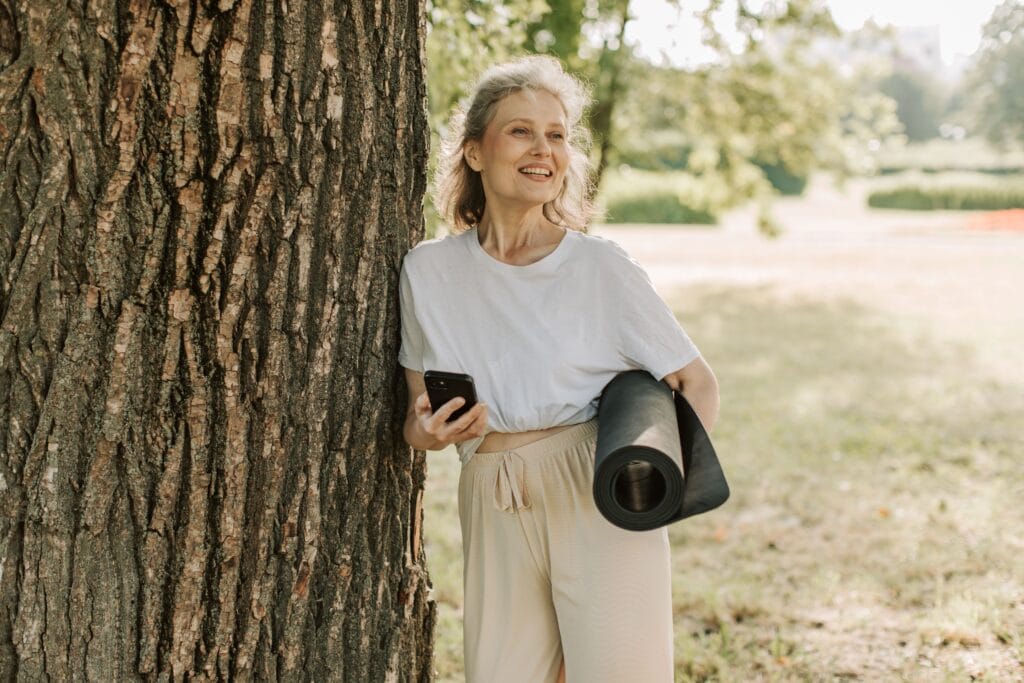 Frau ab 50 lehnt lächelnd an einem Baum mit Yogamatte und Smartphone – aktiv und selbstwirksam im Neuanfang