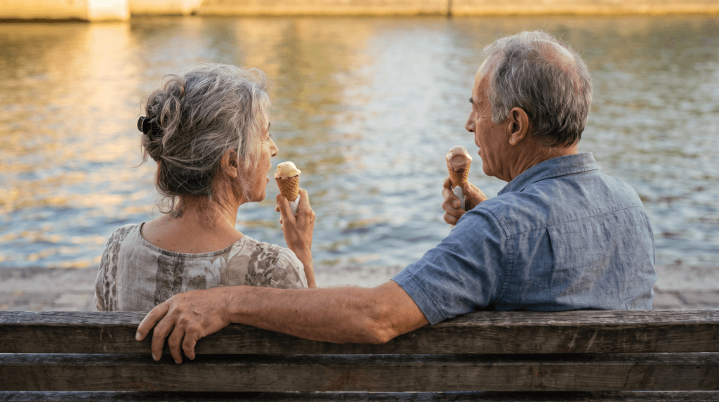 Zwei Menschen ab 60 sitzen mit Eis in der Hand auf einer Bank am Wasser und sind ins Gespräch vertieft